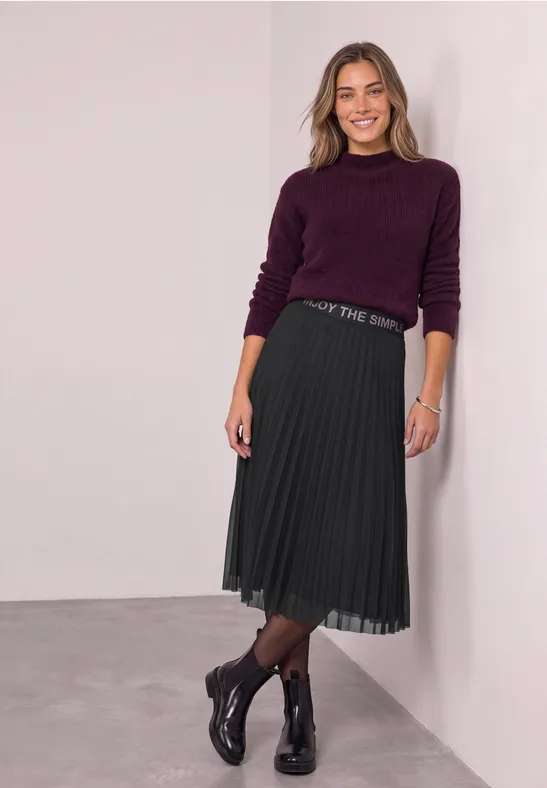 Lächelnde Frau in violettem Pullover, Plissee-Midirock und Stiefeletten lehnt an weißer Wand.