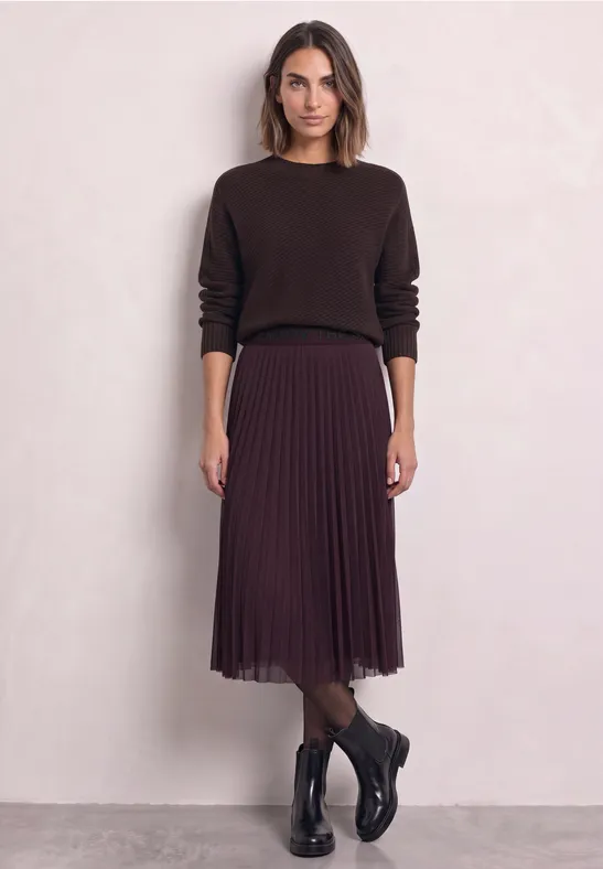 Frau in dunkelbrauem Pullover, plissiertem Midi-Rock und schwarzen Stiefeln vor hellem Hintergrund.