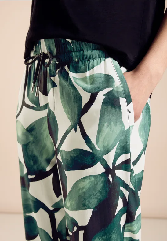 Hose mit grünem Blattmuster und schwarzem Shirt, Hand in der Tasche.