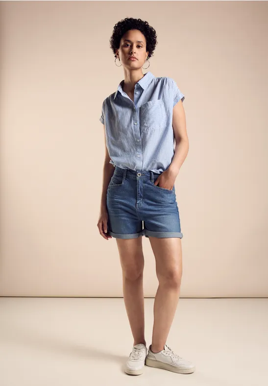 Frau mit kurzen, lockigen Haaren trägt gestreiftes Hemd, Jeansshorts und weiße Sneaker.