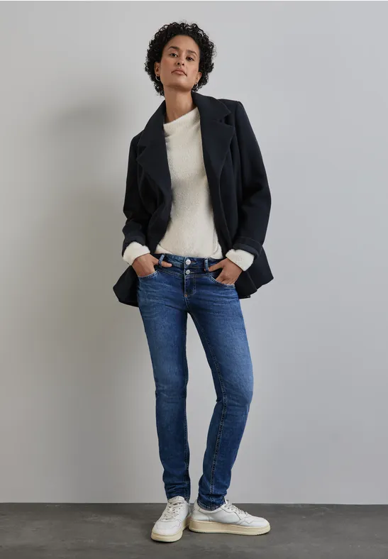 Frau in Jeans, weißem Pullover und dunklem Mantel steht lässig an der Wand.