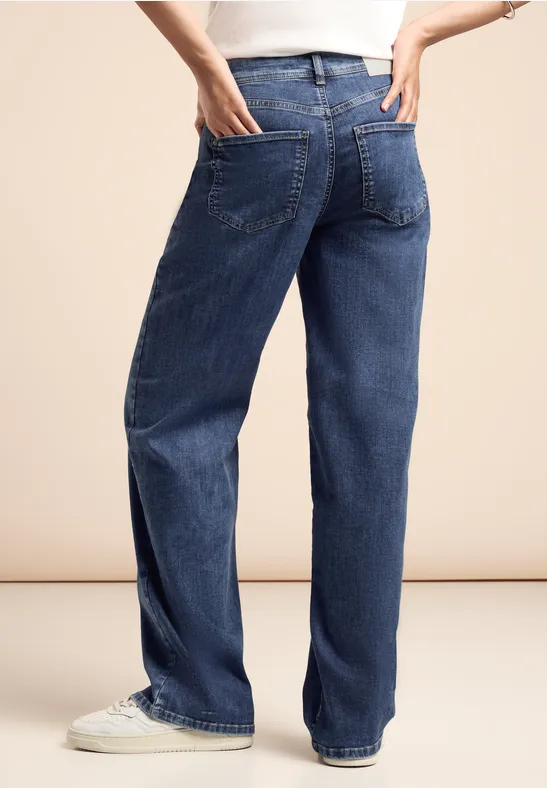 Person trägt blaue Jeans und weiße Sneakers, steht mit Hand in der Gesäßtasche.
