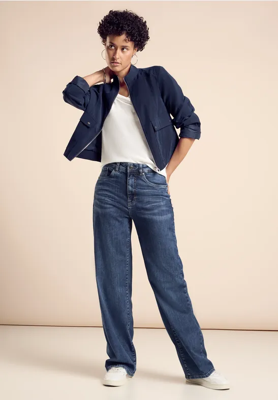 Frau in lässiger Jeans, weißem Shirt, dunkler Jacke und Sneakers vor beigem Hintergrund.