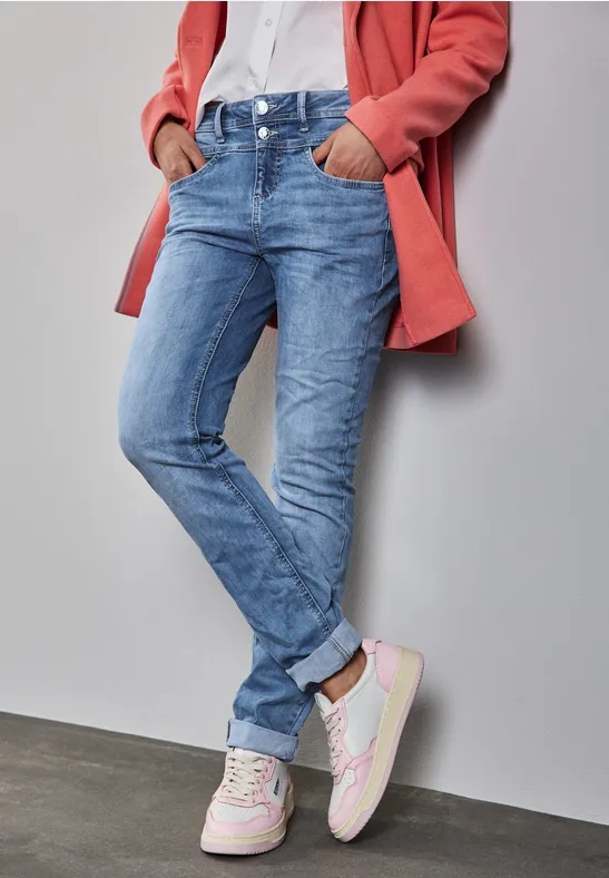 Person in Jeans, weißen Sneakern, weißem Hemd und rosa Mantel lehnt an der Wand.