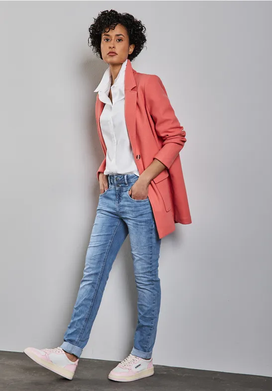 Frau mit rosa Blazer, weißem Hemd, Jeans und Sneakern lehnt an einer Wand.