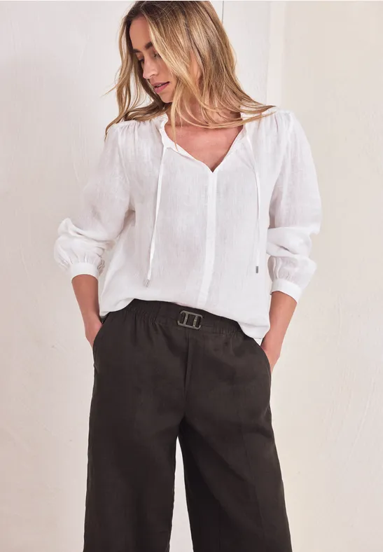 Vrouw in witte blouse en zwarte, wijde broek met riem tegen neutrale achtergrond.