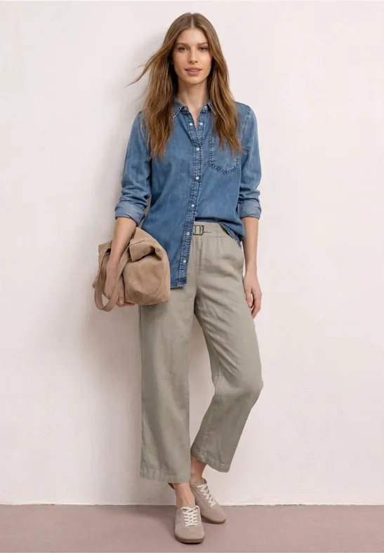 Vrouw in denimblouse, wijde broek, comfortabele schoenen, met een tas en een hoed in de hand.
