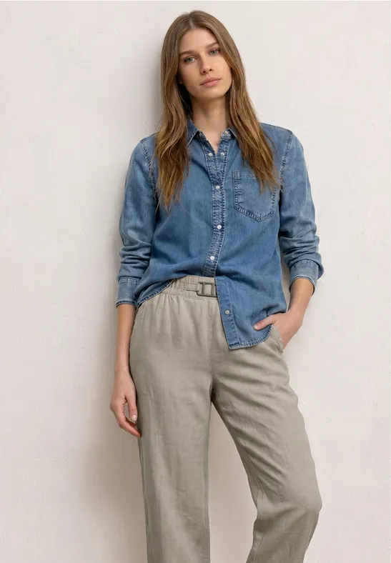 Vrouw draagt een denim blouse en beige stofbroek, staat nonchalant met handen in de zakken.