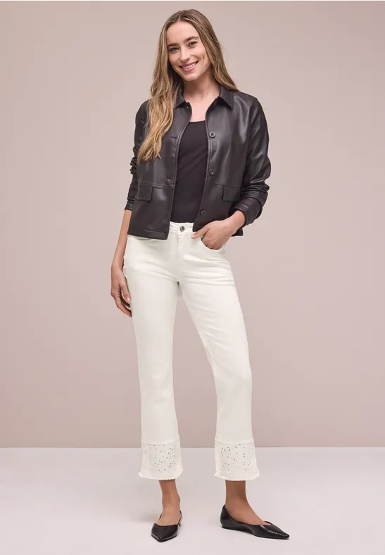 Frau in schwarzer Lederjacke, schwarzer Bluse, weißen Cropped-Hosen mit Spitzen und spitzen schwarzen Ballerinas vor beigem Hintergrund.