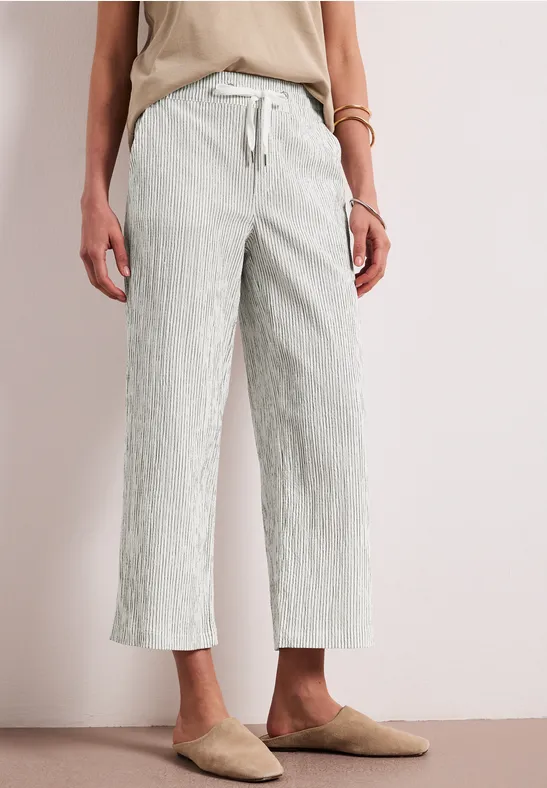 Vrouw draagt crème mules en wit, licht gestreept 7/8 wijde broek, neutrale achtergrond.