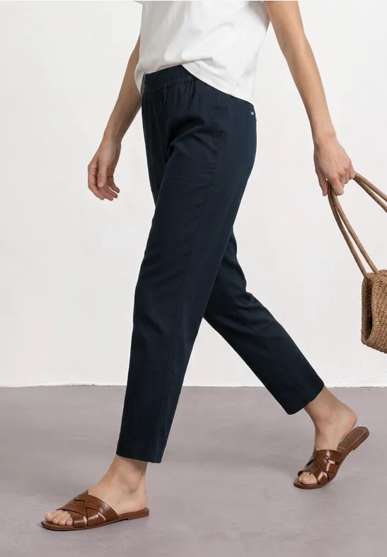 Frau trägt weiße Bluse, dunkelblaue 7/8-Hose, flache Sandalen und hält Korbtasche.