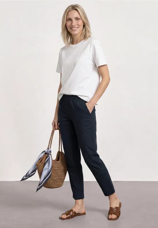 Frau in weißem T-Shirt, dunklen Hosen, geflochtenen Sandalen und Strohtasche mit Tuch.