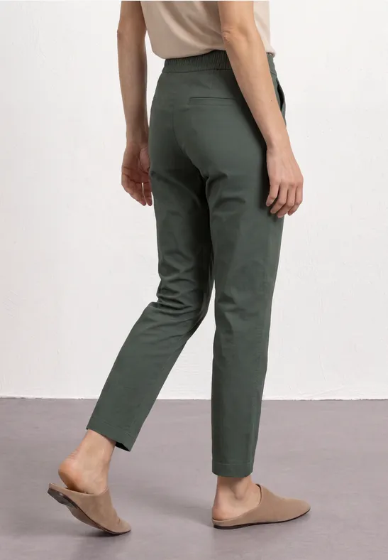 Iemand draagt een groene, slanke broek en comfortabele bruine mules.