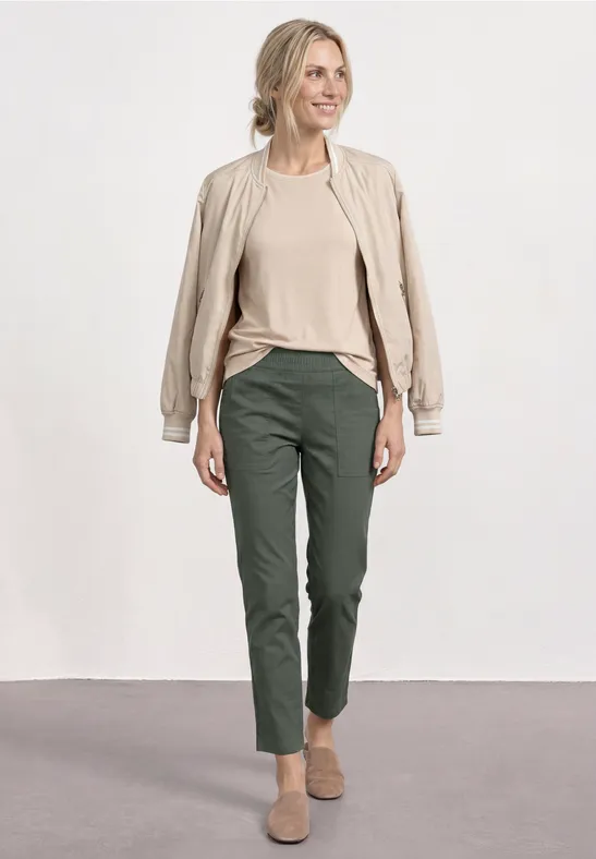 Een vrouw draagt een beige jas, een beige shirt, donkergroene broek en beige schoenen voor een neutrale achtergrond.