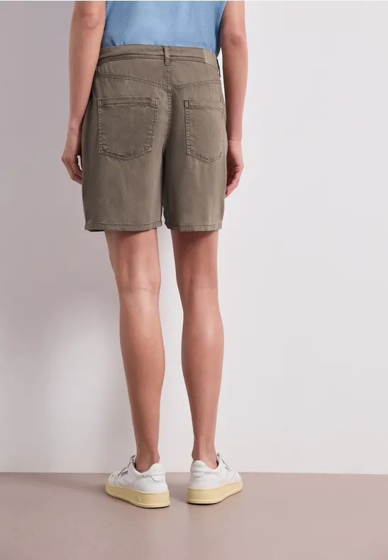 Person von hinten in khakifarbenen Shorts und weißen Turnschuhen, stehend vor neutralem Hintergrund.