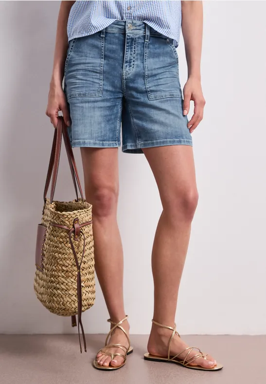 Jambes en short en jean, sac en paille tressée et sandales à lanières dorées.