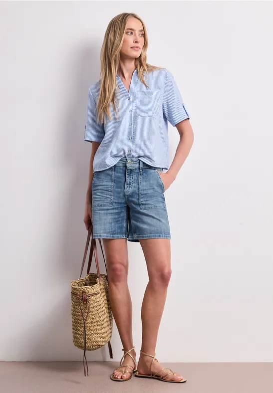 Femme en chemise bleu clair, jupe en jean, sandales, tenant un sac en osier.