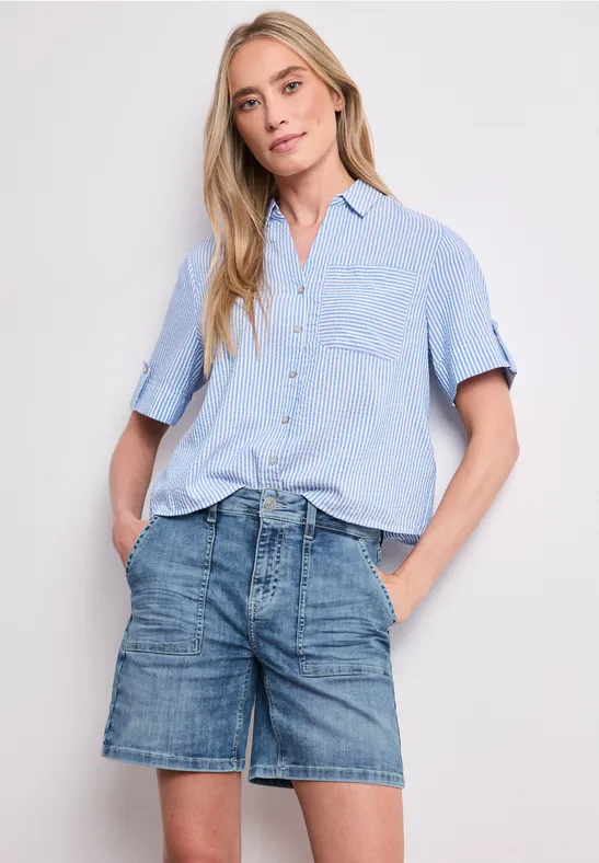 Une femme porte une chemise bleu clair et un short en jean, les mains dans les poches