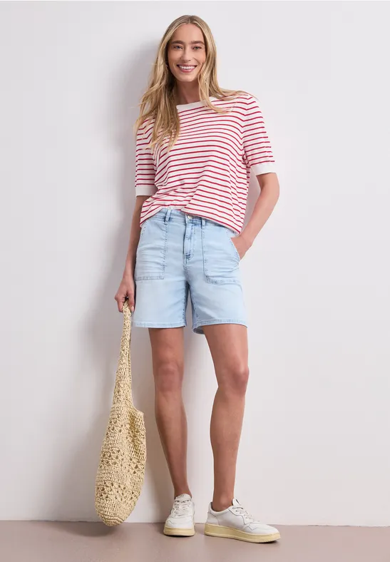 Frau in rot-weiß gestreiftem T-Shirt, hellblauen Jeans-Shorts, Makramee-Tasche und weißen Sneakern.
