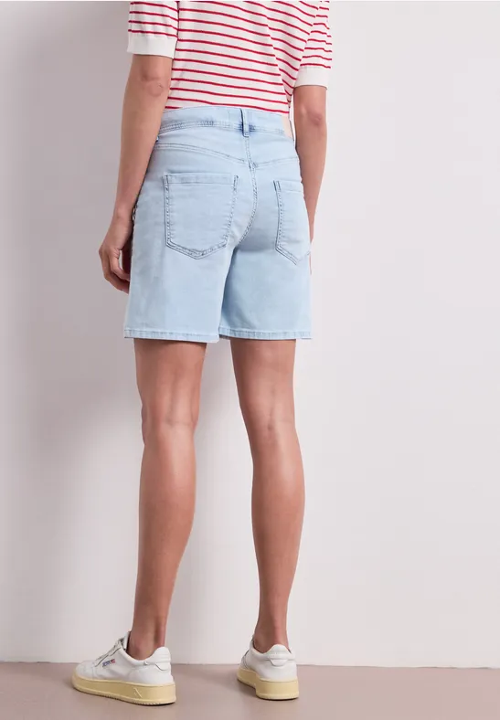 Eine Person steht mit hellblauen Jeansshorts, rotem gestreiften Oberteil und weißen Sneakern vor einer Wand.
