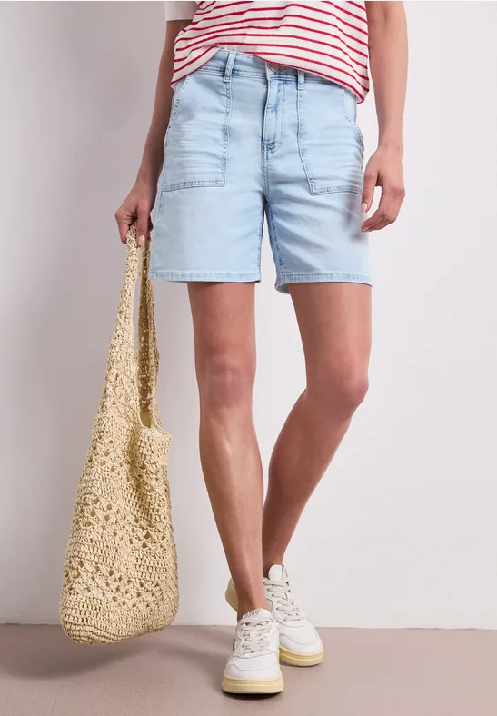 Frau in hellblauen Jeansshorts, gestreiftem Shirt, weißen Sneakern und Häkel-Tasche.