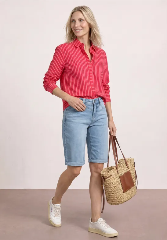 Vrouw in rood hemd, lichtblauwe jeansshorts, witte sneakers en rieten tas.