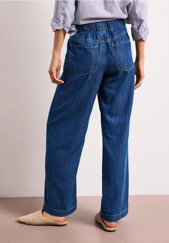 Achteraanzicht van een persoon in wijde blauwe spijkerbroek met elastische tailleband en beige slippers.