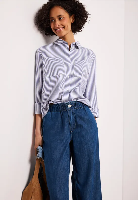 Vrouw met gestreept overhemd, wijde blauwe broek en een tas in de hand.