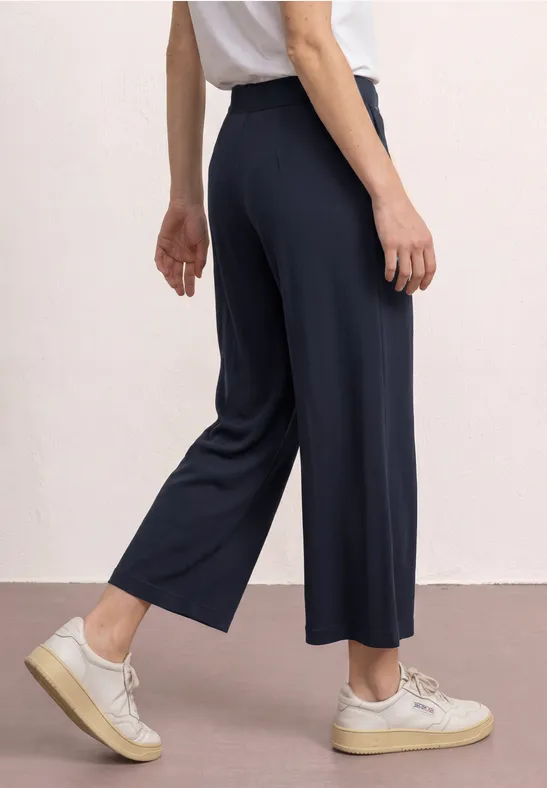 Een persoon draagt wijde, donkerblauwe halflange broek en witte sneakers.