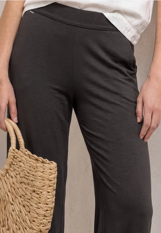 Een persoon draagt wijde donkergrijze broek met elastische taille en houdt een gevlochten strooitas vast.