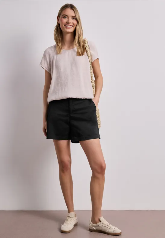 Frau mit weißem T-Shirt, schwarzen Shorts und hellen Sneakers steht vor weißem Hintergrund, Hand in Tasche.