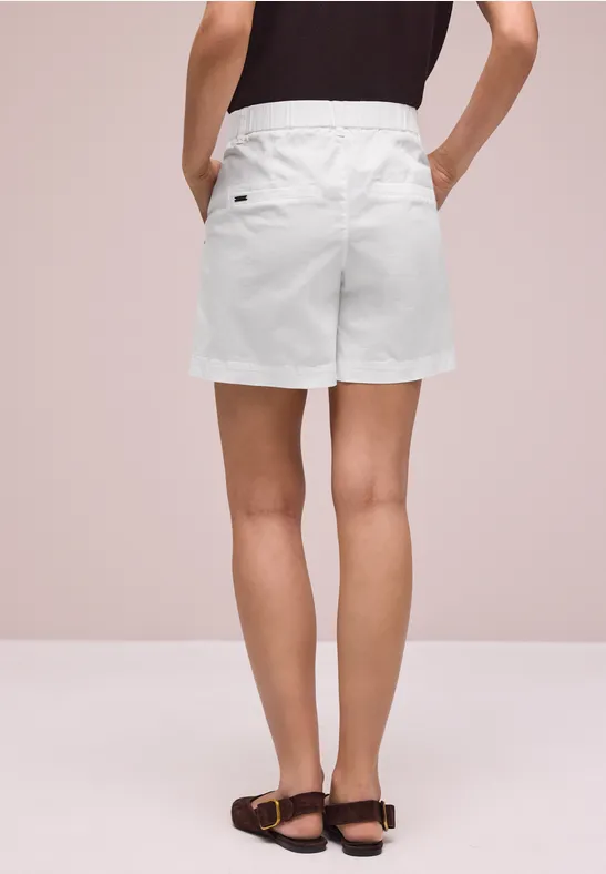 Vrouw van achteren in witte short, zwart top en zwarte sandalen voor een neutrale achtergrond.