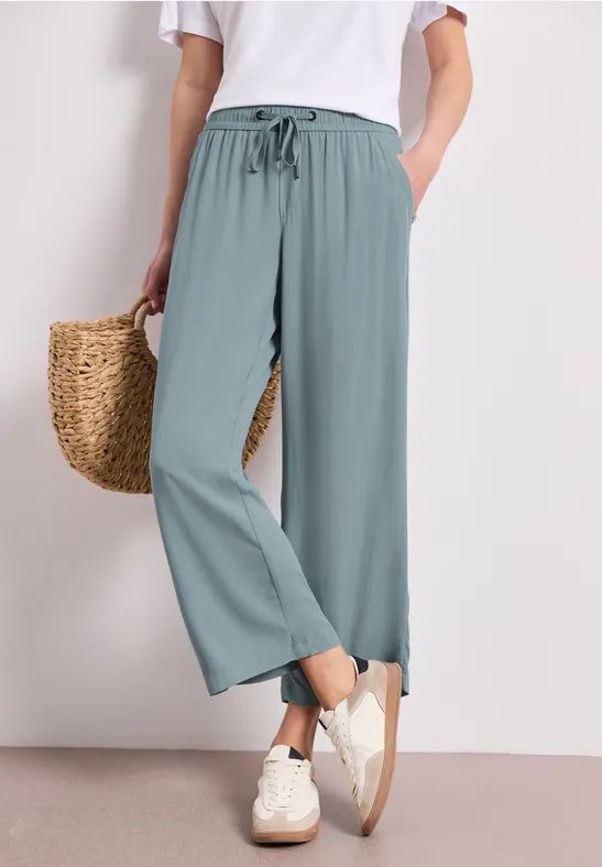 Vrouw draagt lichtblauwe brede broek met elastische taille, wit topje, witte sneakers en een strooien tas.