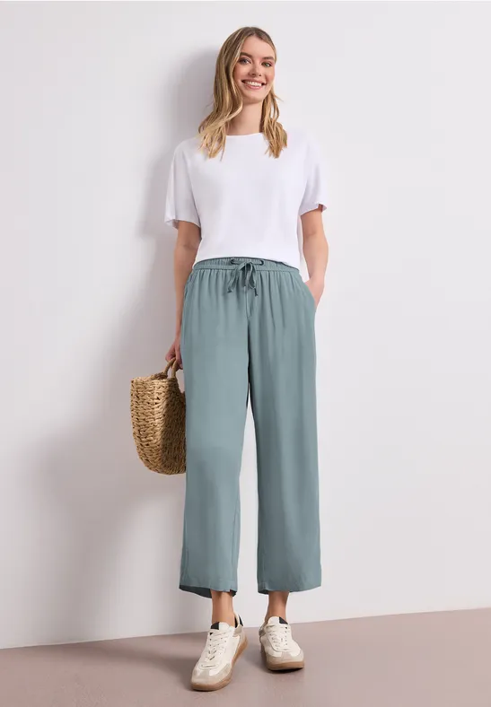 Vrouw in wit T-shirt, losse groene broek en sneakers, met een gevlochten mand.