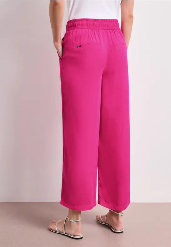 Vue de dos d’une personne portant un pantalon rose fuchsia ample et des sandales blanches sur fond neutre
