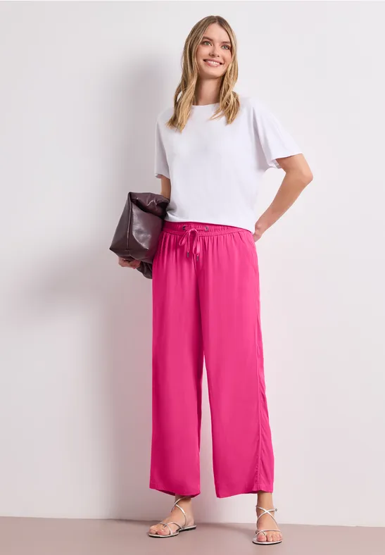 Femme en t-shirt blanc et pantalon large fuchsia, sandales aux pieds et pochette sombre à la main.