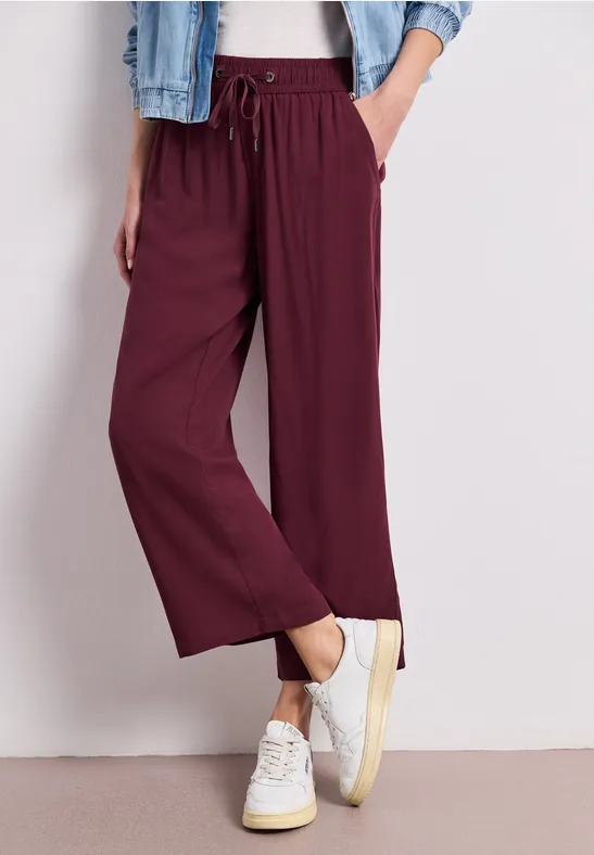 Draagt een wijde bordeaux broek met elastische taille en koord, witte sneakers en een spijkerjas.
