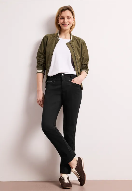 Personne en bomber vert olive, t-shirt blanc, jean noir et mocassins marron, adossée avec décontraction à un mur.
