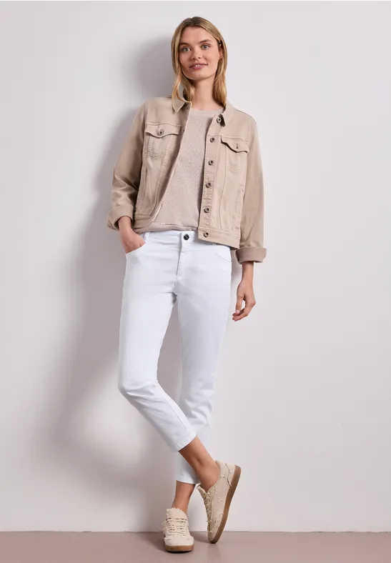 Frau in beiger Jeansjacke, hellbraunem Shirt, weißen Jeans und Espadrilles vor neutralem Hintergrund.