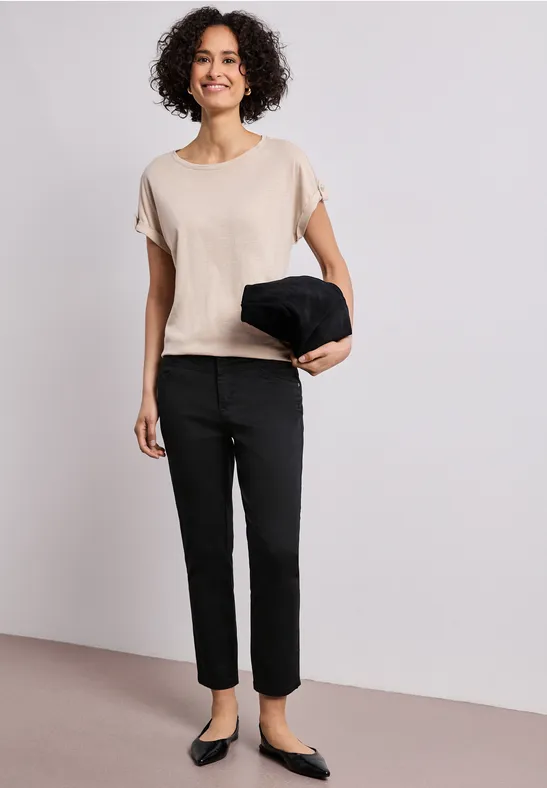 Person in beigem Shirt, schwarzen Hosen und Ballerinas hält eine schwarze Jacke vor neutralem Hintergrund.