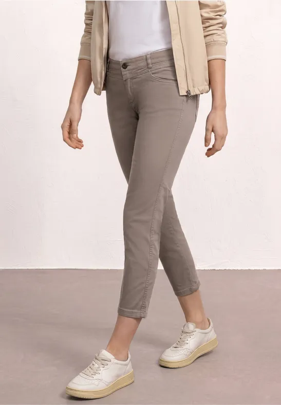 Femme en pantalon slim taupe, baskets blanches, t-shirt blanc et veste beige sur fond neutre.