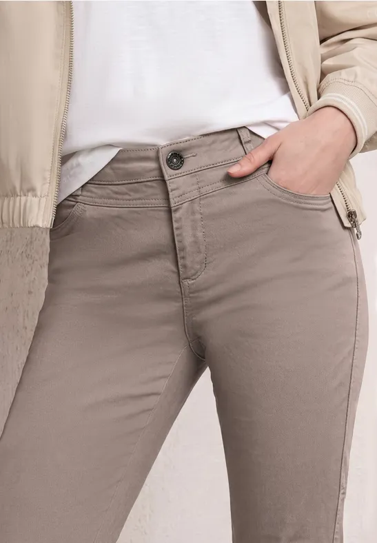 Personne en pantalon taupe, t-shirt blanc et blouson beige clair, main détendue dans la poche.