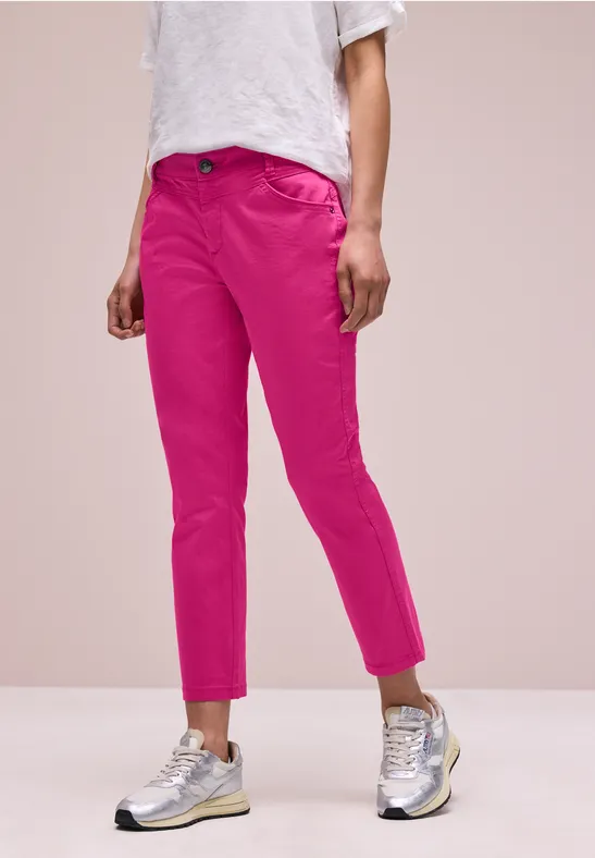 Een persoon draagt felroze, enkelhoge broek, een wit T-shirt en zilverkleurige sneakers voor een roze achtergrond.