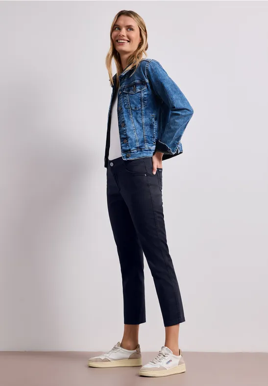 Een vrouw draagt een blauw denimjasje, zwarte broek en witte sneakers, staand voor een lichte achtergrond.