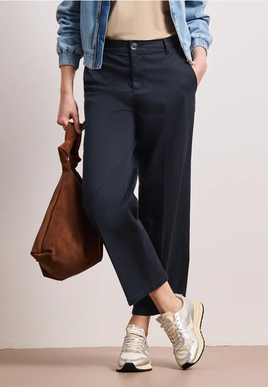 Beinansicht einer Person mit Jeansjacke, beigem Top, dunklen Hosen, Sneakern und brauner Tasche.