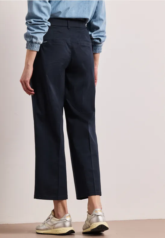 Rückenansicht einer Person in dunkelblauen, geraden Hosen und hellen Sneakers vor hellem Hintergrund.