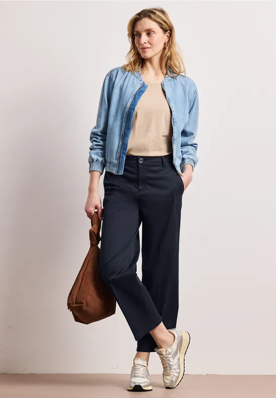 Frau in hellblauer Jacke, beigem Top, schwarzen Hosen und weißen Sneakers mit brauner Tasche.