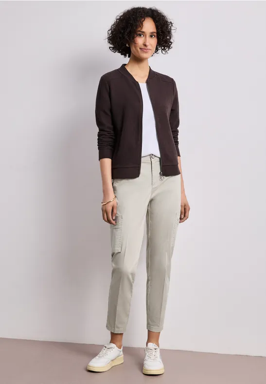 Vrouw met krullend haar in een donker vest, witte top, lichtgekleurde broek en witte sneakers tegen een witte achtergrond.