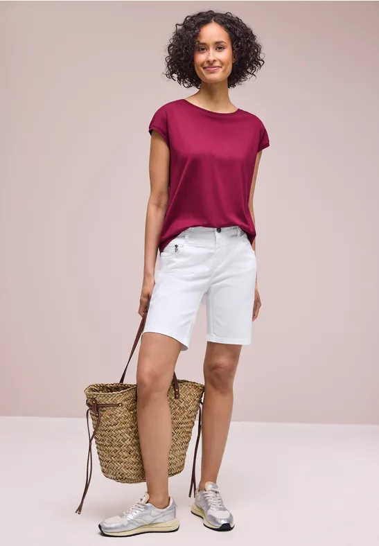 Een vrouw draagt een bordeauxrood T-shirt, witte shorts en sneakers, met een gevlochten strandtas.