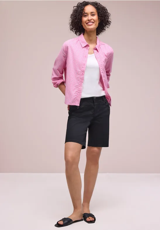 Femme portant une chemise rose, un t-shirt blanc, un short bermuda noir et des sandales noires sur fond clair.